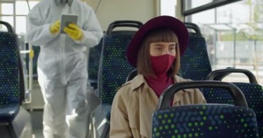 Koruyucu giysili ve maskeli bir sağlık görevlisi otobüsteki kadın yolcuları tarıyor. Sıcaklık kontrolü yaparken toplu taşımada oturan genç bir kadın. Covid-19 Konsepti.
