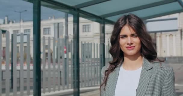 Attraente ragazza bruna sorridente e passeggiando vicino a un moderno edificio per uffici. Giovane bella strada millenaria in posa e guardando la fotocamera in strada della città. Il concetto di stile di vita . — Video Stock