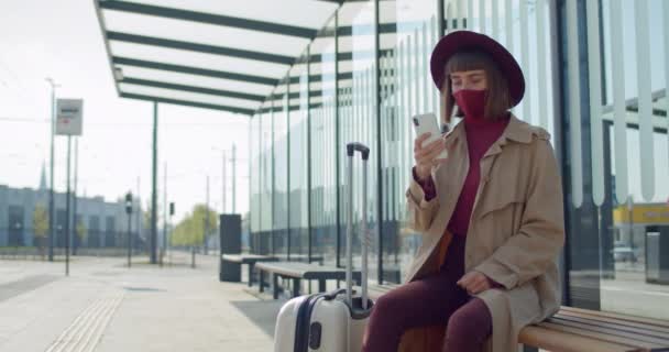 Dziewczyna w bawełnianej masce protekcyjnej za pomocą smartfona podczas siedzenia na ławce w pobliżu lotniska. Podróżująca kobieta w stylowych ubraniach z walizką patrząc na ekran telefonu. Pojęcie koronawiru. — Wideo stockowe