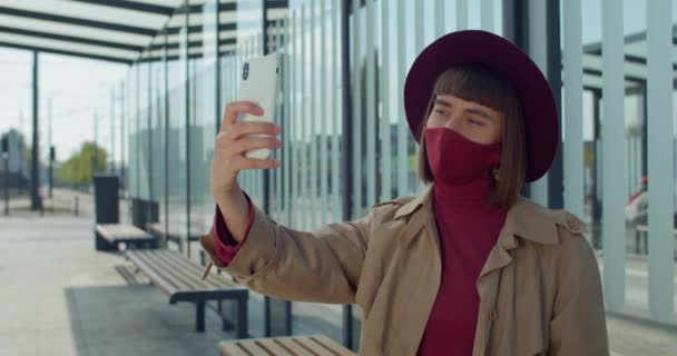 Mujer joven en máscara de algodón tomando selfies cerca de edificio de vidrio moderno. Atractiva chica millennial posando mientras usa la cámara frontal del teléfono inteligente. Concepto de protección contra el coronavirus . — Vídeo de stock