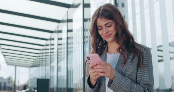 Millenniial veselá žena se dívá na obrazovku telefonu a psaní zpráv na ulici. Atraktivní brunetka dívka s úsměvem při použití smartphone a hlazení vlasů v blízkosti moderní budovy. — Stock video
