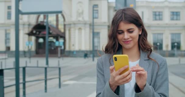 街の通りを歩いている間、陽気な若い女性のスクロールとタッチスクリーンのクロップビュー。彼女のスマートフォンを使って魅力的な女の子をブルネットと笑顔でニュースフィードをチェックします。屋外. — ストック動画