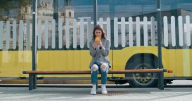 Neşeli kadın günlük giysiler giyip internete takılıyor ya da sosyal ağlarda sohbet ediyor. Güzel kız toplu taşıma durağında otururken akıllı telefon kullanıyor. Arka planda otobüs yolculuğu.