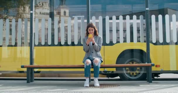 Χαρούμενη γυναίκα που φοράει καθημερινά ρούχα που περιηγείται στο διαδίκτυο ή κουβεντιάζει στα κοινωνικά δίκτυα. Όμορφο κορίτσι που χρησιμοποιεί το smartphone, ενώ κάθεται σε στάση μέσα μαζικής μεταφοράς. Βόλτα με λεωφορείο στο παρασκήνιο. — Αρχείο Βίντεο
