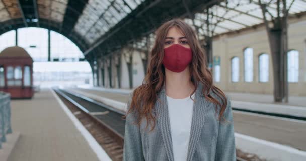 Porträtt av ung kvinna i bomull ansiktsmask tittar på kameran på tom järnvägsstation. Beskär utsikt över smillennial brunett flicka stående ensam. Begreppet virus och pandemi. — Stockvideo