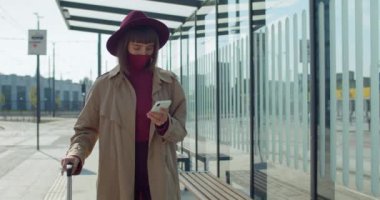 Havaalanı yakınlarında akıllı telefon kullanan koruyucu maskeli ve valizli şık bir kız. Milenyum kadın yolcusu yürüyor ve telefon ekranına bakıyor. Koronavirüs ve seyahat kavramı.