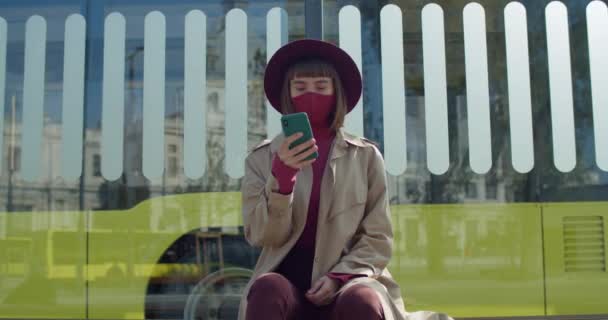 Vista de la cosecha de la mujer con estilo utilizando el teléfono inteligente mientras está sentado en la parada de transporte público. Chica en la máscara protectora de algodón mirando la pantalla del teléfono mientras se desplaza noticias. Autobús en segundo plano . — Vídeos de Stock