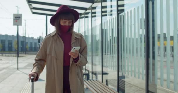 空港近くのスマートフォンを使用して保護マスクやスーツケースのスタイリッシュな女の子。ミレニアル世代の女性の乗客の歩行や携帯電話の画面を見て。コロナウイルスの概念と旅行. — ストック動画