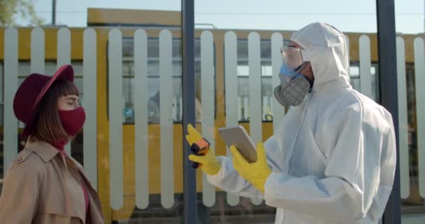 Travailleur médical en combinaison de protection et masque tenant le comprimé et filtrant le passager féminin. La jeune femme a de la fièvre tout en vérifiant la température à la gare. Concept de Covid-19 . — Video