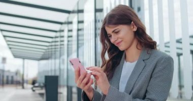 Çekici genç bir kadının telefon ekranını kaydırması ve şaşırmış yüz ifadesi. Milenyum esmer kız internette geziniyor ve modern ofisin yanında otururken gülümsüyor..