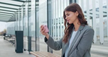 Çekici milenyum kızı yatay manzara modunda akıllı telefonu tutarken ve ekrana bakarken yan görüntüsü. Genç bir kadın bankta otururken video izleyip gülümsüyor. Açık havada..