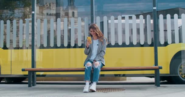 Młoda kobieta korzystająca ze smartfona podczas siadania na przystanku komunikacji publicznej.Millenijna dziewczyna w ubraniach przeglądająca internet lub czatująca w sieciach społecznościowych. Autobus w tle. — Wideo stockowe