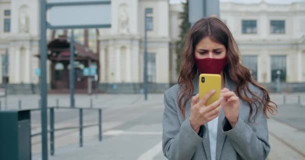 Junge Frau mit Gesichtsmaske checkt die neuesten Nachrichten oder surft im Internet. Millennial Girl in wiederverwendbarer Baumwollmaske mit Smartphone und Scrollbildschirm beim Spazierengehen auf einer leeren Stadtstraße. — Stockvideo
