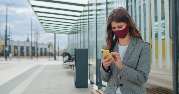 Brunette girl in cotton mask checking news or browsing internet. Millennial woman in protection handmade face mask using smartphone and scrolling screen while sitting at bus stop.