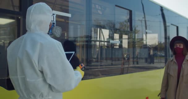 Medische medewerker in beschermende pak screent passagiers voor de instapbus. Jonge vrouwelijke reiziger met koffer met temperatuurcontrole. Concept van bescherming tegen het coronavirus en reizen. — Stockvideo