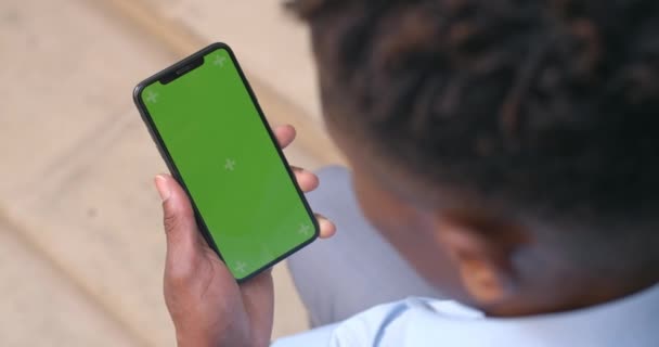 Sopra la spalla vista di ragazzo afro americano seduto e guardando lo schermo verde del telefono con punti di tracciamento. L'uomo che tiene in mano uno smartphone finto. Concetto di chiave cromatica. All'aperto . — Video Stock