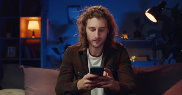 Jeune beau mec avec des cheveux longs naviguant sur Internet tout en étant assis sur le canapé le soir. Homme hipster millénaire avec des boucles d'oreilles en utilisant un smartphone et en regardant l'écran à la maison . — Video