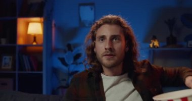 Yakışıklı genç sakallı adamın elinde patlamış mısır kasesiyle kanepede oturup televizyonu açmasını yakından izleyin. Milenyum yakışıklı adam televizyon izliyor ve uzaktan kumanda kullanıyor..