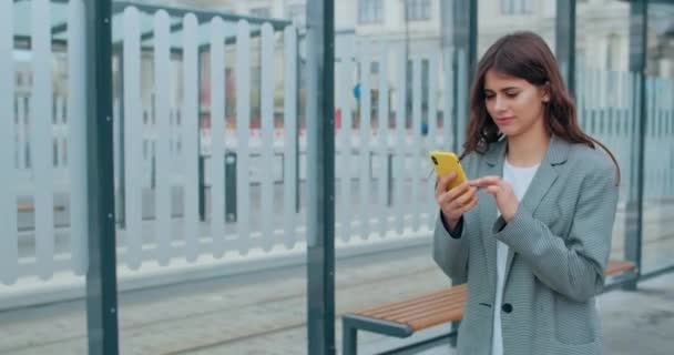 Tysiącletnie brunetka kobieta przewijanie, dotykanie ekranu podczas przeglądania Internetu. Ładna dziewczyna korzystająca ze smartfona i uśmiechnięta podczas spaceru po nowoczesnej ulicy miasta. Pojęcie stylu życia. — Wideo stockowe