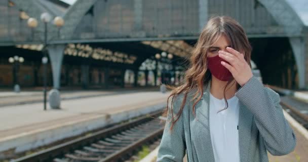 Portrait of young woman in cotton facial mask standing at railway station. Crop view of serious millennial girl looking to camera and strocking hair. Concept of virus pandemic. — Stock Video