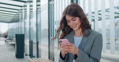 Genç iş kadını internette geziniyor ve modern ofis merkezinin yanında otururken telefon ekranına bakıyor. Sohbet etmek için akıllı telefonu kullanırken gülümseyen mutlu milenyum kızları.