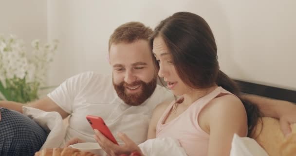 Meglepett férfi és nő nézi a telefon képernyőn, és mosolyog, miközben fekszik az ágyon. Vidám romantikus pár használ okostelefon csodálkozó arcát reggeli közben. A szabadidő fogalma. — Stock videók