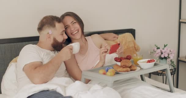 Mutlu genç çift telefon ekranına bakarken konuşuyor ve gülümsüyor. Kadın yatakta kahvaltıda akıllı telefon tutarken erkek kahve içiyor. İlişki ve eğlence kavramı. — Stok video