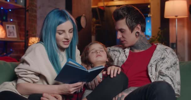 Retrato de la joven familia moderna pasando tiempo junto con el libro en la noche. Hermosa mamá leyendo en voz alta mientras su marido con tatuajes y túneles escuchando y besando a su hija dormida . — Vídeos de Stock