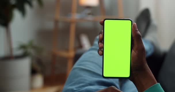Afrikai férfi fekszik a kanapén hamis képernyős okostelefonnal a kezében. A fickó telefonál, miközben a zöld képernyőt nézi és üzenetet olvas. A krómkulcs fogalma. Hangulatos otthoni háttér. — Stock videók