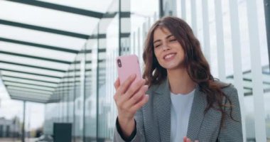 Çekici kız görüntülü konuşma yaparken gülümsüyor ve konuşuyor. Milenyum iş kadını el sallarken modern bina yakınlarında iletişim kurmak için akıllı telefon uygulaması kullanıyor..