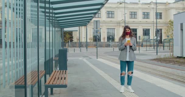 Giovane donna elegante indossa abiti casual con maschera riutilizzabile in cotone sul suo viso guardando alla fotocamera. Ragazza millenaria in possesso di smartphone e in piedi alla fermata del trasporto pubblico . — Video Stock