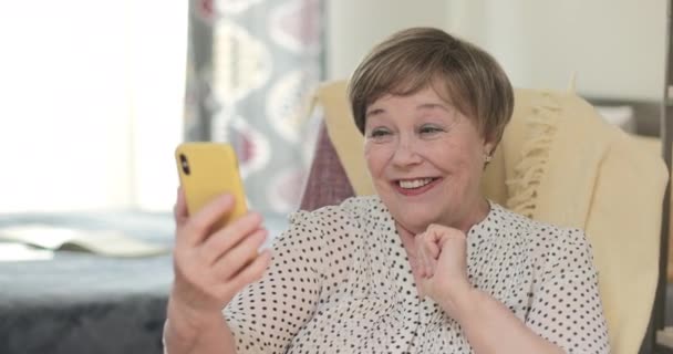 Happy grandmother in 60s having video call on her modern smartphone. Cheerful elderly woman talking, blowing kiss and waving during video conversation. Concept of communication. — Stock Video