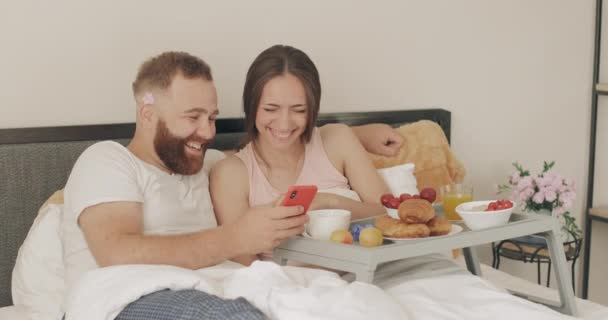 Happy young couple spending time and laughing in bed. Man and woman looking at smartphone screen and talking while having breakfast in early morning. Concept of leisure and relationship. — Stock Video