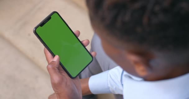 彼の手に緑の画面でスマートフォンを保持し、座っているアフロアメリカのビジネスマンの肩のビューで。クロマキーとモックアップの概念。屋外. — ストック動画