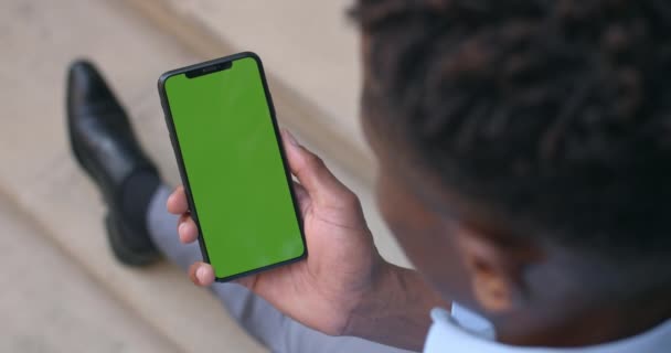 Uomo d'affari afro-americano con smartphone in mano con finto schermo mentre seduto sulle scale.Vista sulla spalla del ragazzo che utilizza il telefono verde. Concetto di chiave cromatica. All'aperto . — Video Stock