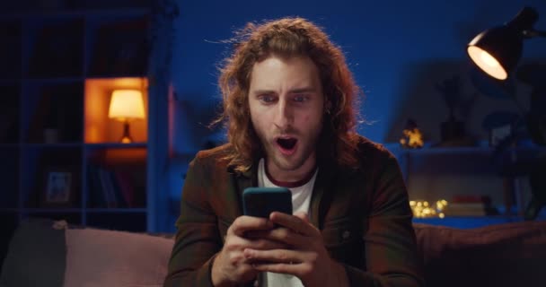 Glada hipster kille gör förvånad ansikte medan du tittar på telefonskärmen. Stilig långhårig man ler och visar emotoner när du använder smartphone på soffan i mysigt hem. — Stockvideo