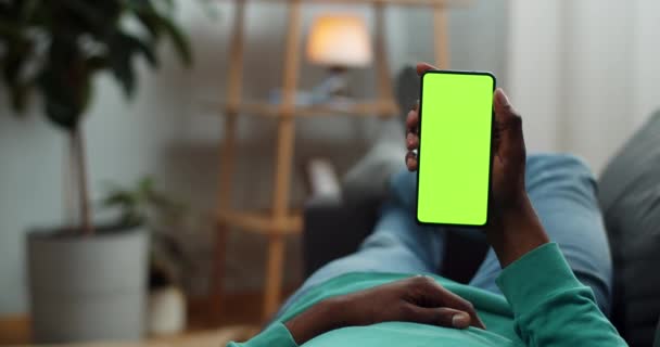 Joven tumbado en el sofá con un moderno teléfono inteligente de pantalla verde en la mano. Chico viendo vídeo mientras mira la pantalla simulada. Concepto de croma key. Acogedor fondo casero . — Vídeo de stock