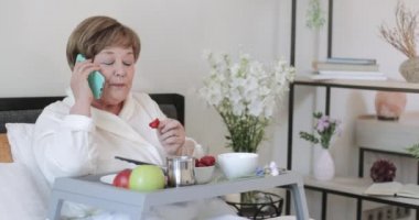 Yatakta kahvaltı yaparken konuşan ve çilek yiyen güzel bir yaşlı kadın. Yaşlı bir kadın, bacaklarında yemek ile otururken akıllı telefondan iletişim kuruyor..