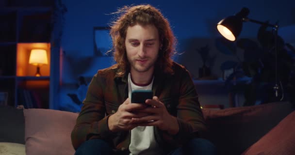 Joven barbudo alegre sonriendo mientras juega. Guapo chico de pelo largo con pendientes usando teléfono inteligente y mirando a la pantalla mientras está sentado en el sofá en la sala de noche . — Vídeos de Stock