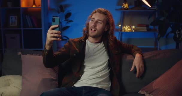 Millenijny bloger uśmiechający się i mówiący siedząc na kanapie w domu. Wesoły facet gestykulujący, mówiący i patrzący na kamerę smartfona podczas rozmowy wideo. Pojęcie stylu życia. — Wideo stockowe