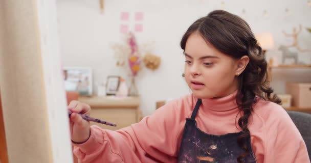 Vista frontal de la hermosa joven sonriendo mientras pinta la imagen en la habitación. Simpática adolescente con síndrome de Down sentado frente a Molbert mientras tiene terapia de arte . — Vídeos de Stock