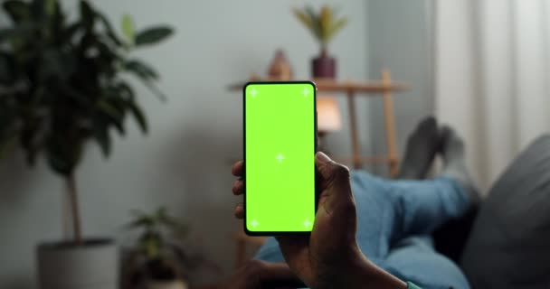 Afro american man holding mobile phone in one hand and looking at mock up screen with trecking markers. Guy lying on sofa and watching video while using smartphone. Concept of chroma key. — Stock Video