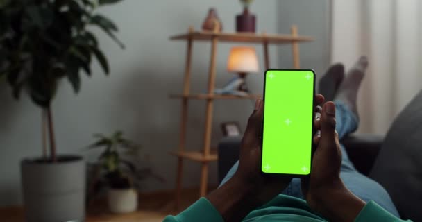 Hombre joven acostado en el sofá con pantalla verde y marcadores de trecking smartphone en sus manos. Guy leyendo noticias o libro electrónico mientras mira la pantalla simulada. Concepto de croma key. Acogedor fondo casero . — Vídeos de Stock