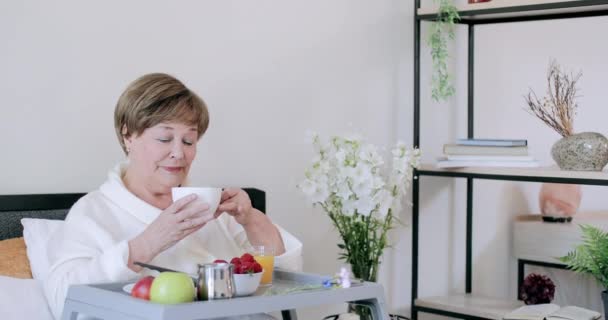 Hermosa mujer jubilada que le ordena su café de la mañana mientras desayuna en la cama. Vieja mujer guapa en Rob sosteniendo la taza y oliéndola mientras está sentada con Trey lleno de comida . — Vídeos de Stock