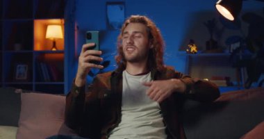 Happy young man recording video message for friend and sending while sitting on sofa at home. Handsome hipster guy talking and having video call while using smartphone camera.