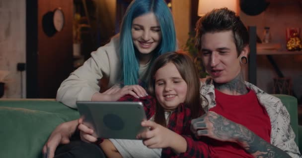 Família moderna feliz assistindo vídeo engraçado ao usar tablet em casa. Jovens pais e sua filha adolescente olhando para exibição de gadget enquanto se sentam no sofá e passam tempo juntos . — Vídeo de Stock