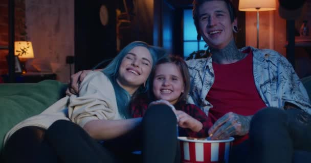 Alegre familia sentada en el sofá y viendo películas divertidas. Pareja moderna e hija adolescente comiendo palomitas de maíz y sonriendo mientras mira la pantalla de televisión. Personas informales con tatuaje y piercing . — Vídeos de Stock