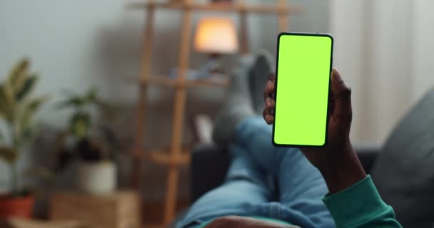 Um afro-americano deitado no sofá e a segurar um telemóvel moderno. Homem lendo e livro enquanto olha para a tela verde. Conceito de chave croma e simular. Aconchegante casa fundo . — Vídeo de Stock