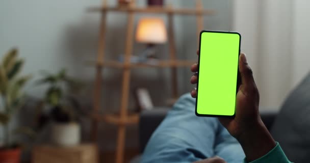 Hombre afroamericano acostado en un sofá con un moderno teléfono de pantalla verde en la mano. Chico leyendo las últimas novedades mientras busca pantalla simulada. Concepto de croma key. Acogedor fondo casero . — Vídeo de stock