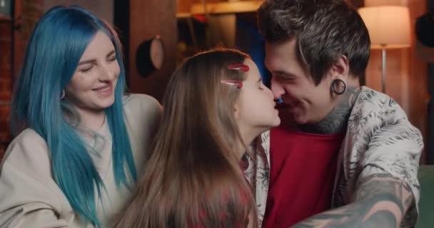 Happy father with tattoos and ear tunnels rubbing noses with his daughter while sitting with blue haired mom on sofa. Portrait of cheerful modern family having fun at home together. — Wideo stockowe